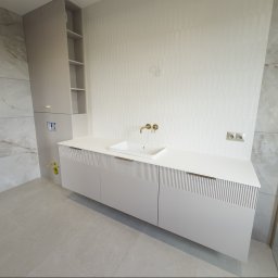 I WOOD DESIGN - Jasna łazienka z szafką pod umywalkę z frezowanymi frontami, złotą armaturą i wysoką szafką z półkami. Na ścianach płytki imitujące marmur i biała mozaika.