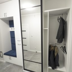 I WOOD DESIGN - Biała szafa wnękowa z przesuwnymi drzwiami lustrzanymi i czarnymi ramami, uzupełniona siedziskiem z niebieską tapicerką i szafkami. Widoczne ubrania na wieszakach.