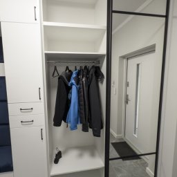 I WOOD DESIGN - Biała szafa wnękowa z czarnymi detalami i lustrem, w przedpokoju. Widoczne ubrania na wieszakach i półki. Minimalistyczny design, szare płytki na podłodze.