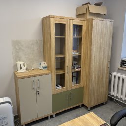 I WOOD DESIGN - Zestaw mebli biurowych z jasnego drewna: szafka z blatem, przeszklona witryna i wysoka szafa. Na blacie czajnik i pudełka. W tle grzejnik i okno z żaluzjami.