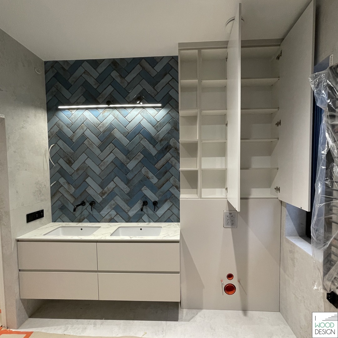 Nowoczesna łazienka z szafką pod umywalkę z dwiema umywalkami, niebieską ścianą z płytek ułożonych w jodełkę i białą szafką z otwartymi półkami.