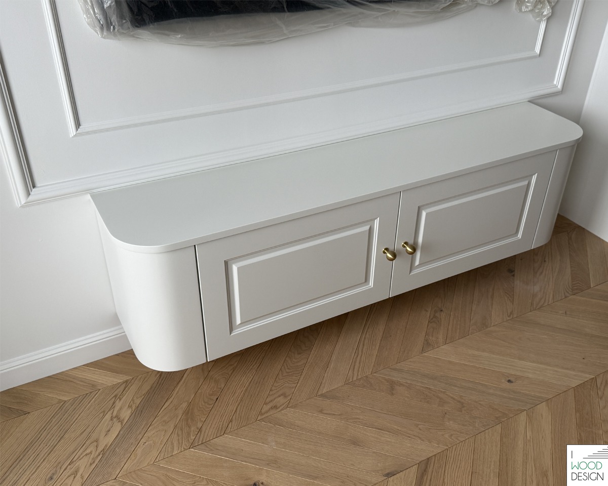 Biała, podwieszana szafka RTV z zaokrąglonym bokiem i złotymi uchwytami na tle ściany z dekoracyjną sztukaterią i parkietu w jodełkę.