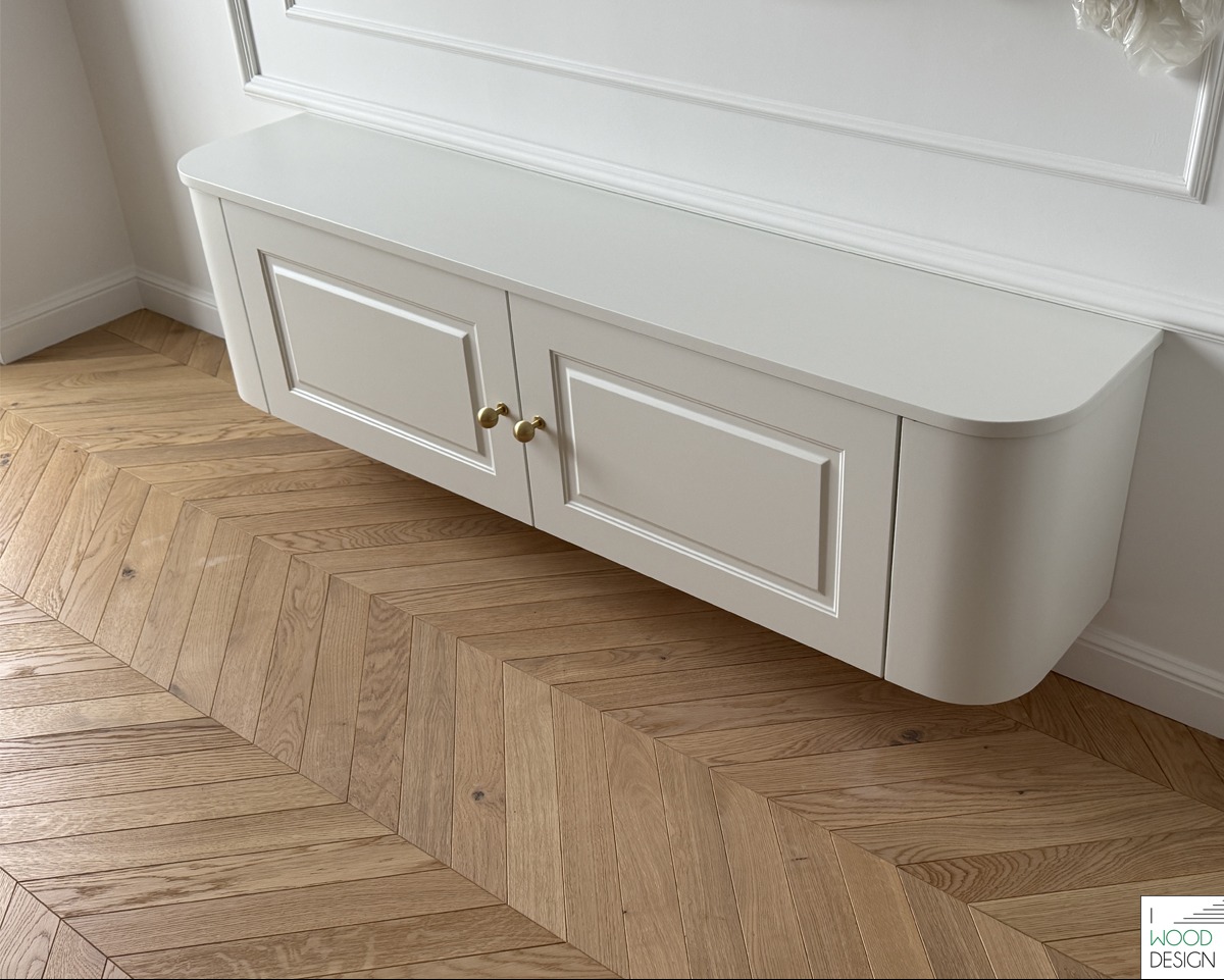 Biała, podwieszana szafka z frezowanymi drzwiczkami i złotymi gałkami na tle parkietu ułożonego we wzór jodełki oraz białej ściany z dekoracyjną listwą.