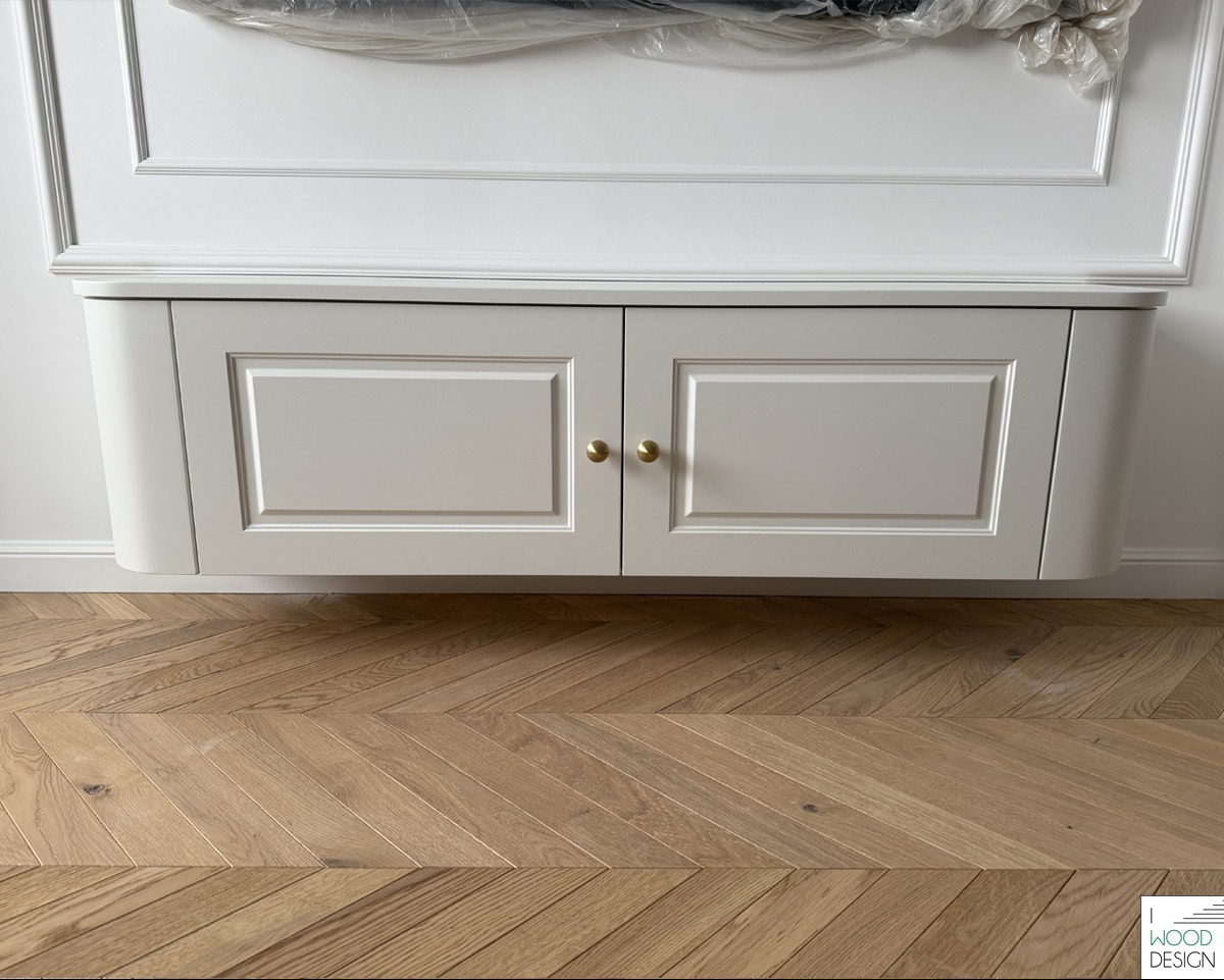 Elegancka, wisząca szafka RTV w jasnym odcieniu, z frezowanymi frontami i złotymi uchwytami, umieszczona na tle białej ściany z dekoracyjną sztukaterią i parkietem w jodełkę.