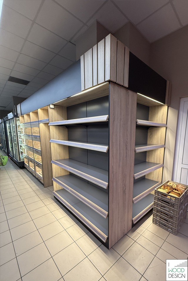 Regał sklepowy z oświetleniem LED i drewnianym wykończeniem, eksponujący pieczywo w skrzynce. Widoczny fragment chłodziarek w tle.