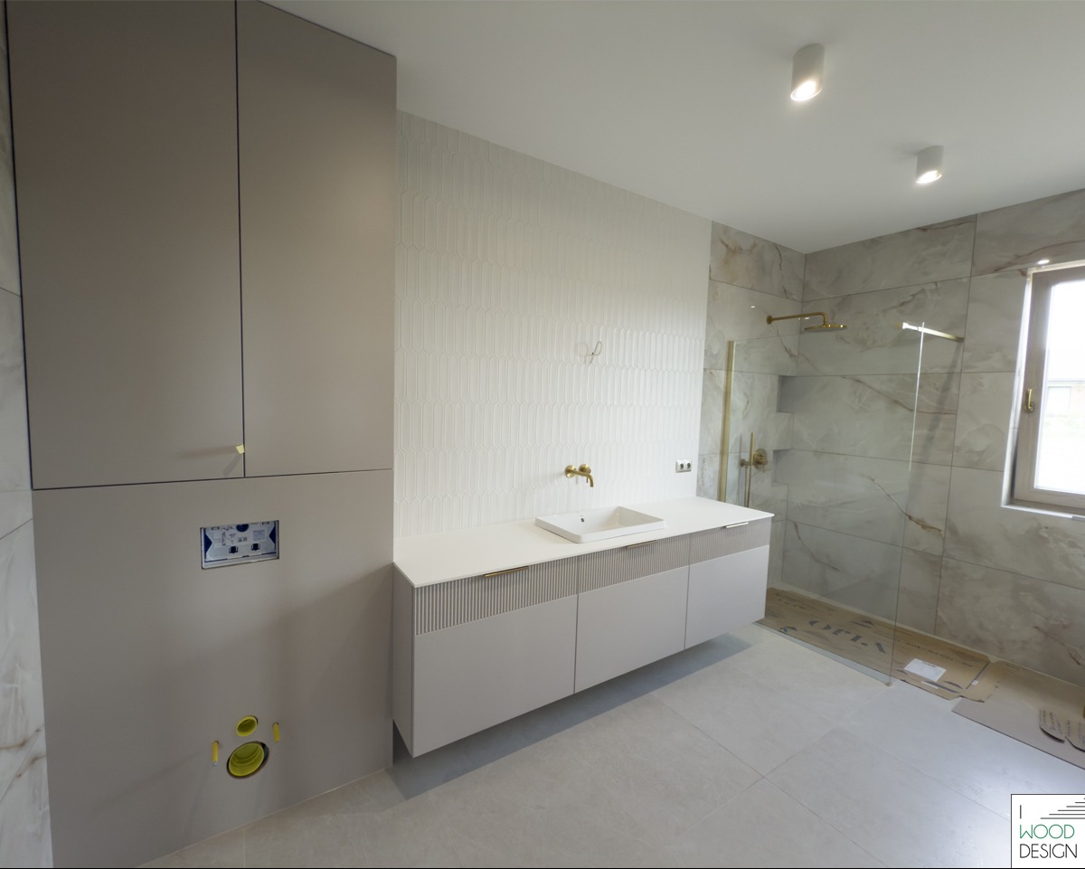 Nowoczesna łazienka z beżowymi szafkami na wymiar, złotą armaturą i kabiną prysznicową z marmurowymi płytkami. Minimalistyczny design z dbałością o detale.