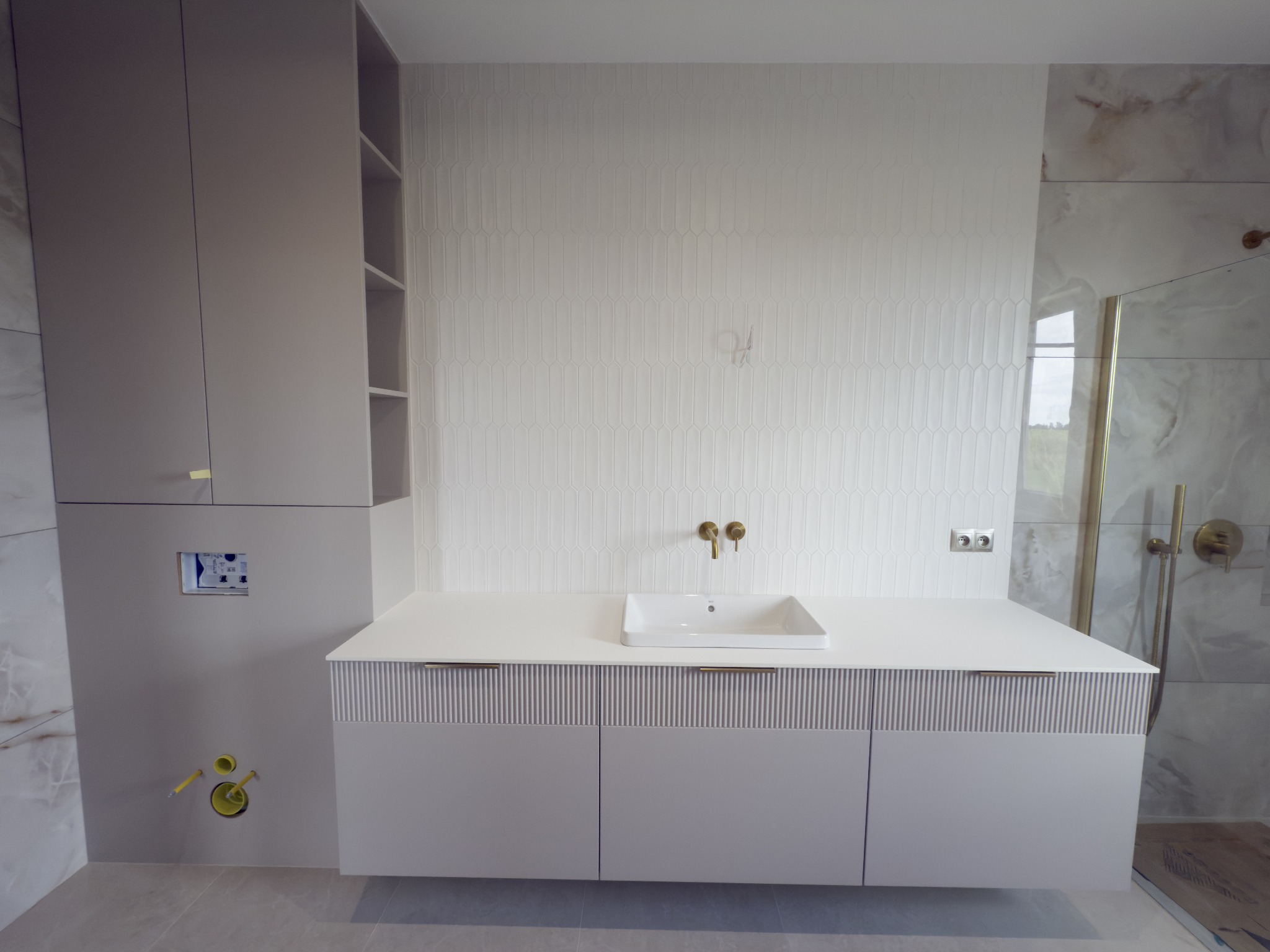 Jasna łazienka z szafką pod umywalką z frezowanymi frontami, złotą armaturą i płytkami imitującymi marmur w strefie prysznica. Nowoczesny design.