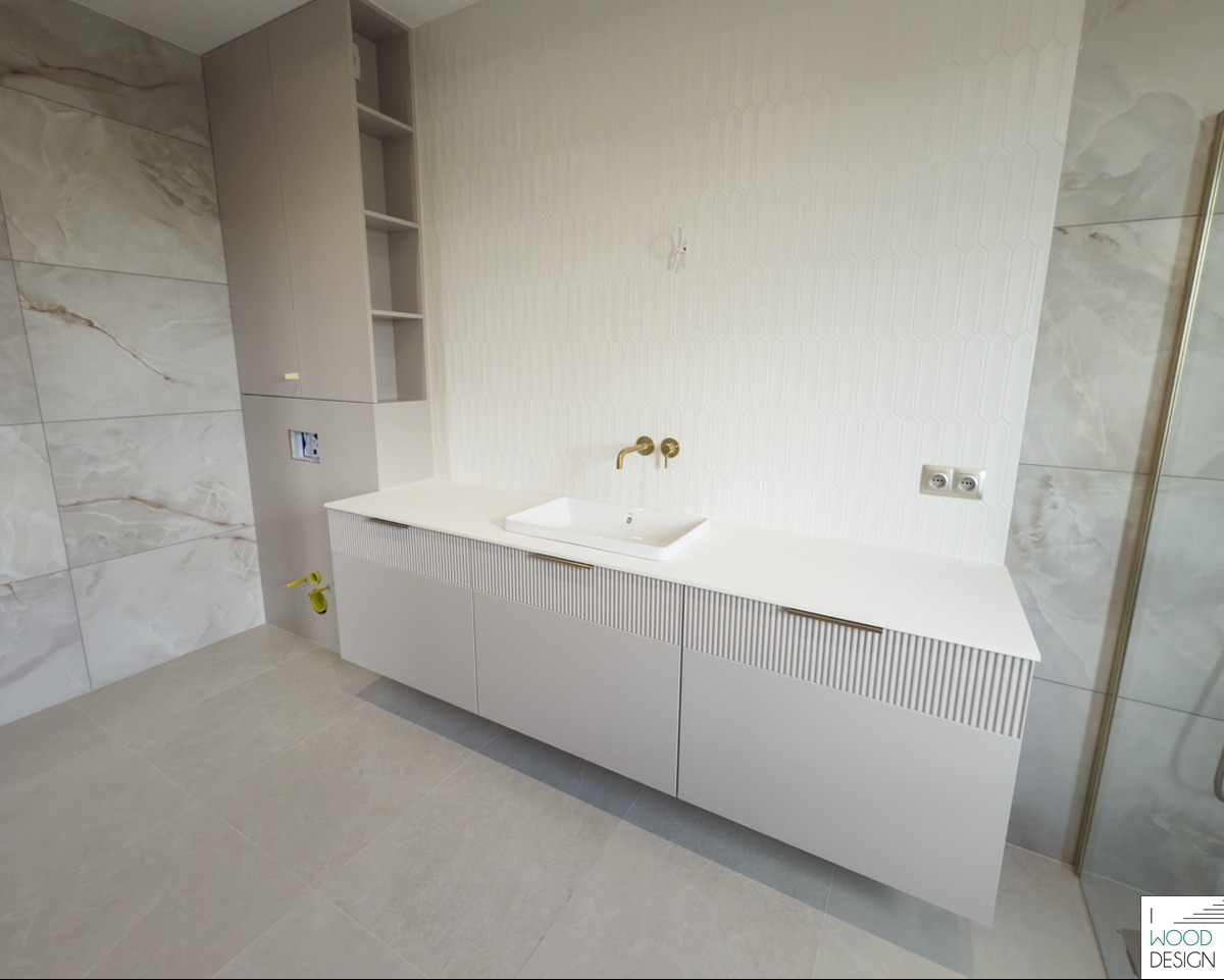 Jasna łazienka z szafką pod umywalkę z frezowanymi frontami, złotą armaturą i wysoką szafką z półkami. Na ścianach płytki imitujące marmur i biała mozaika.