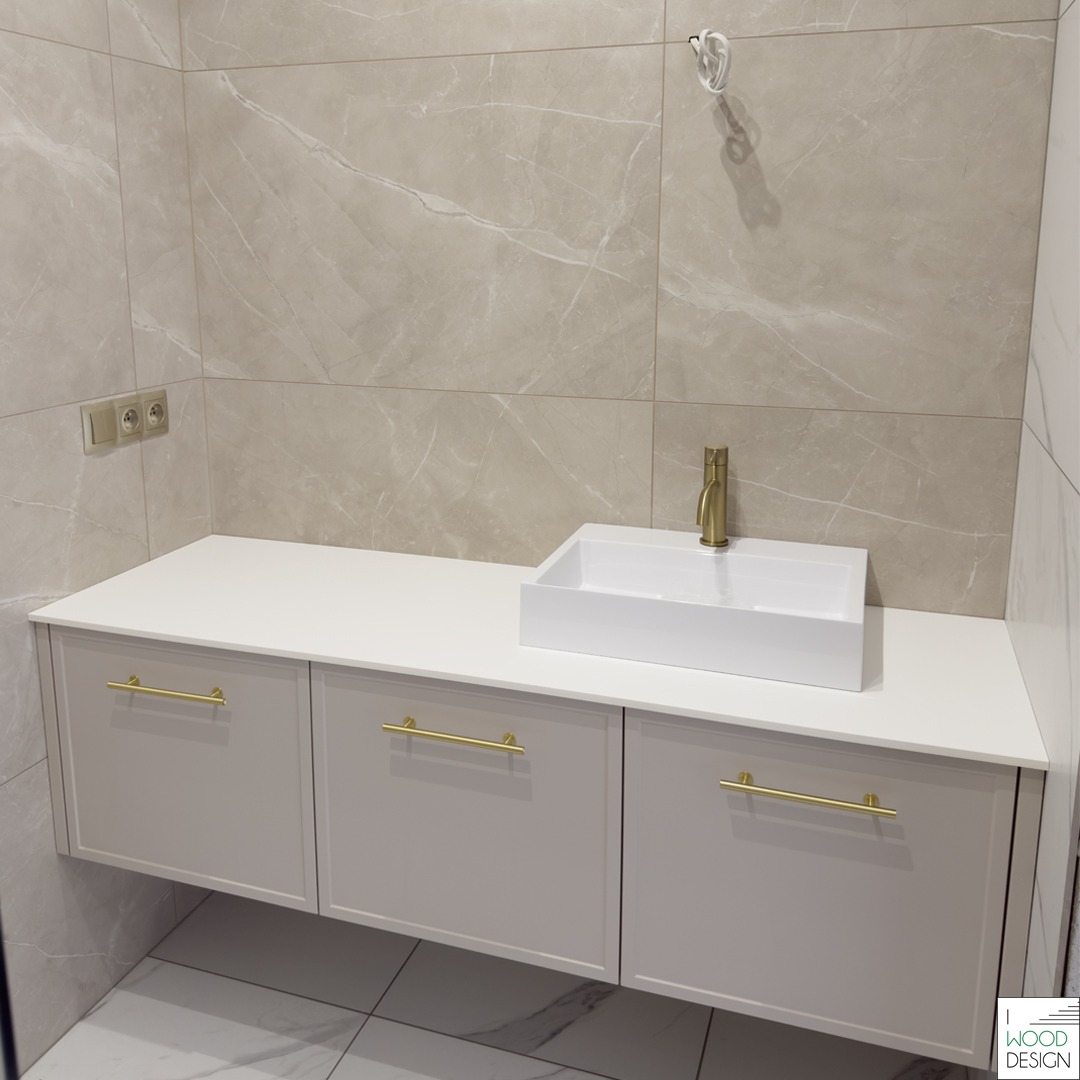 Elegancka szafka łazienkowa pod umywalkę z białym blatem, złotymi uchwytami i nowoczesną umywalką nablatową na tle beżowych płytek imitujących marmur.