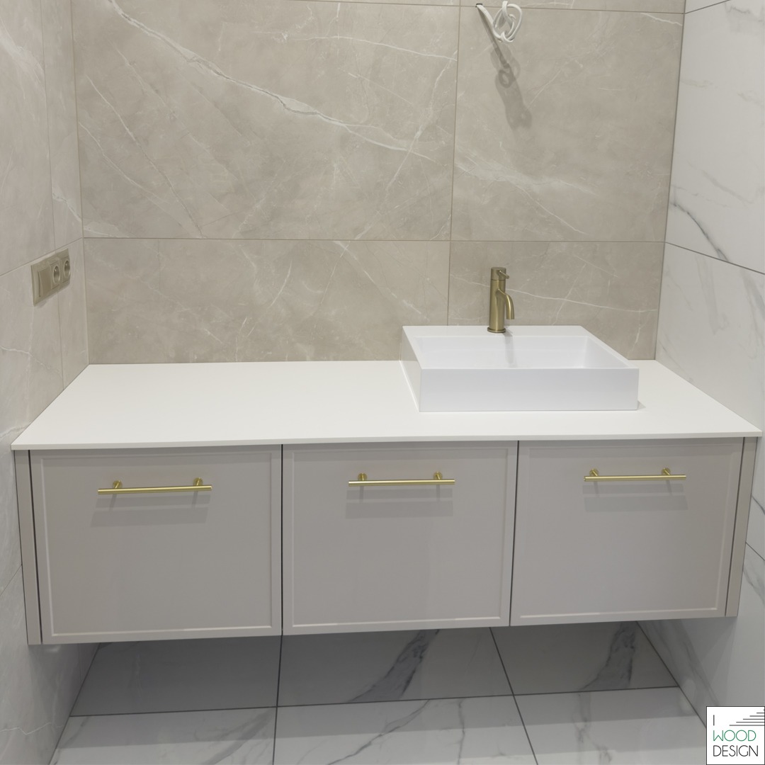 Nowoczesna łazienka z białymi szafkami pod umywalką i złotymi uchwytami, na tle marmurowych płytek. Minimalistyczny design, ciepłe oświetlenie.