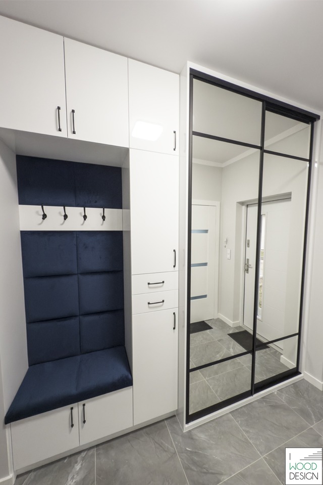Biała szafa wnękowa z siedziskiem i lustrzanymi drzwiami przesuwnymi w przedpokoju. Granatowe obicie siedziska i panelu ściennego. Minimalistyczny design.
