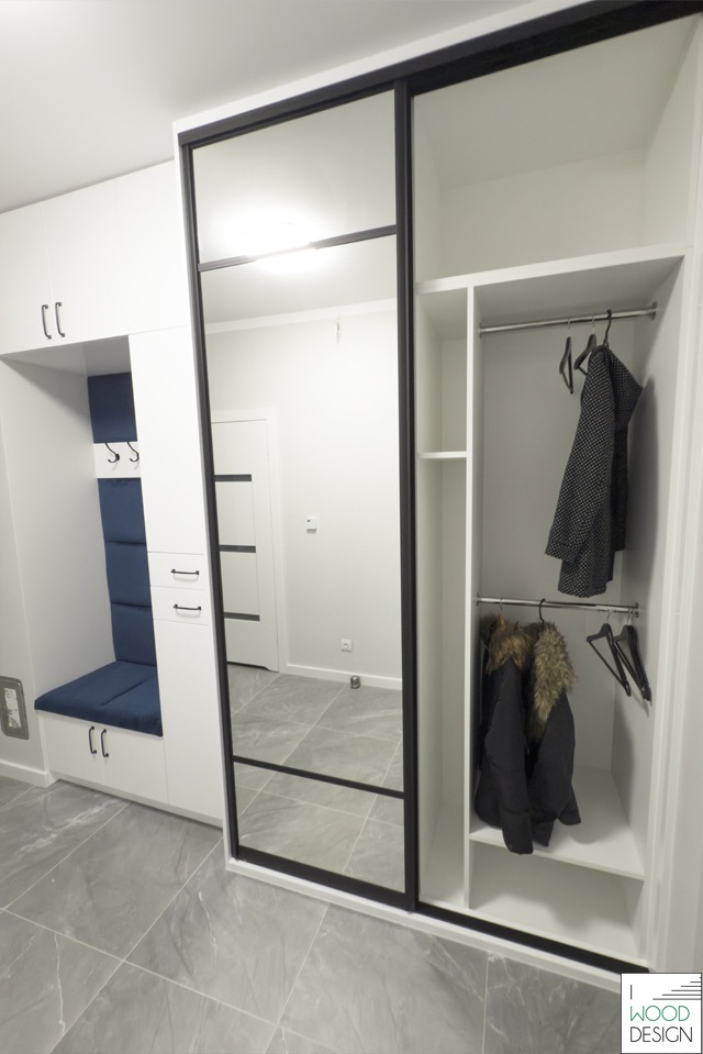 Biała szafa wnękowa z przesuwnymi drzwiami lustrzanymi i czarnymi ramami, uzupełniona siedziskiem z niebieską tapicerką i szafkami. Widoczne ubrania na wieszakach.