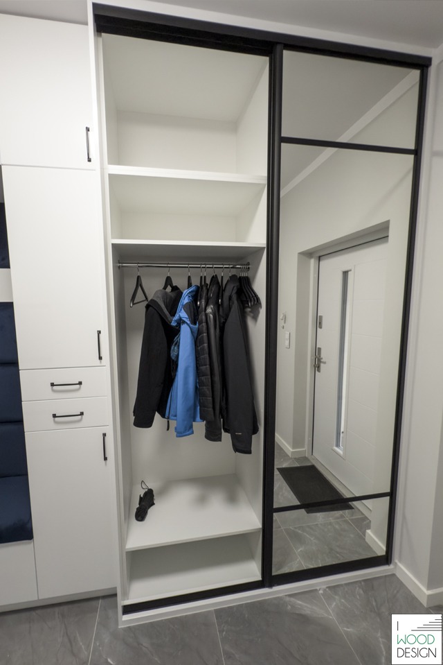 Biała szafa wnękowa z czarnymi detalami i lustrem, w przedpokoju. Widoczne ubrania na wieszakach i półki. Minimalistyczny design, szare płytki na podłodze.