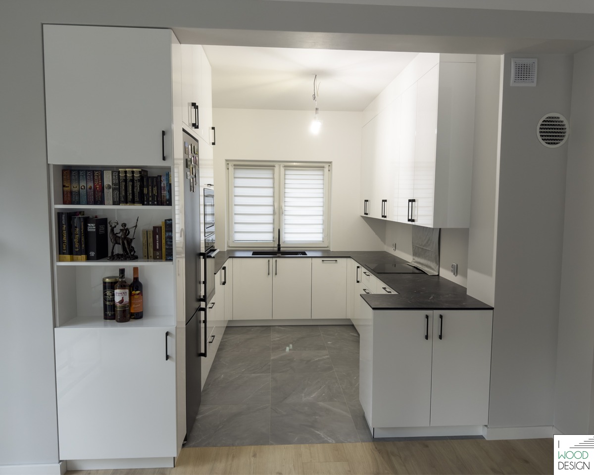 Biała kuchnia z czarnymi blatami i uchwytami, widok z salonu. Na półce książki i butelki. Minimalistyczny design, szare płytki podłogowe, nowoczesne oświetlenie.