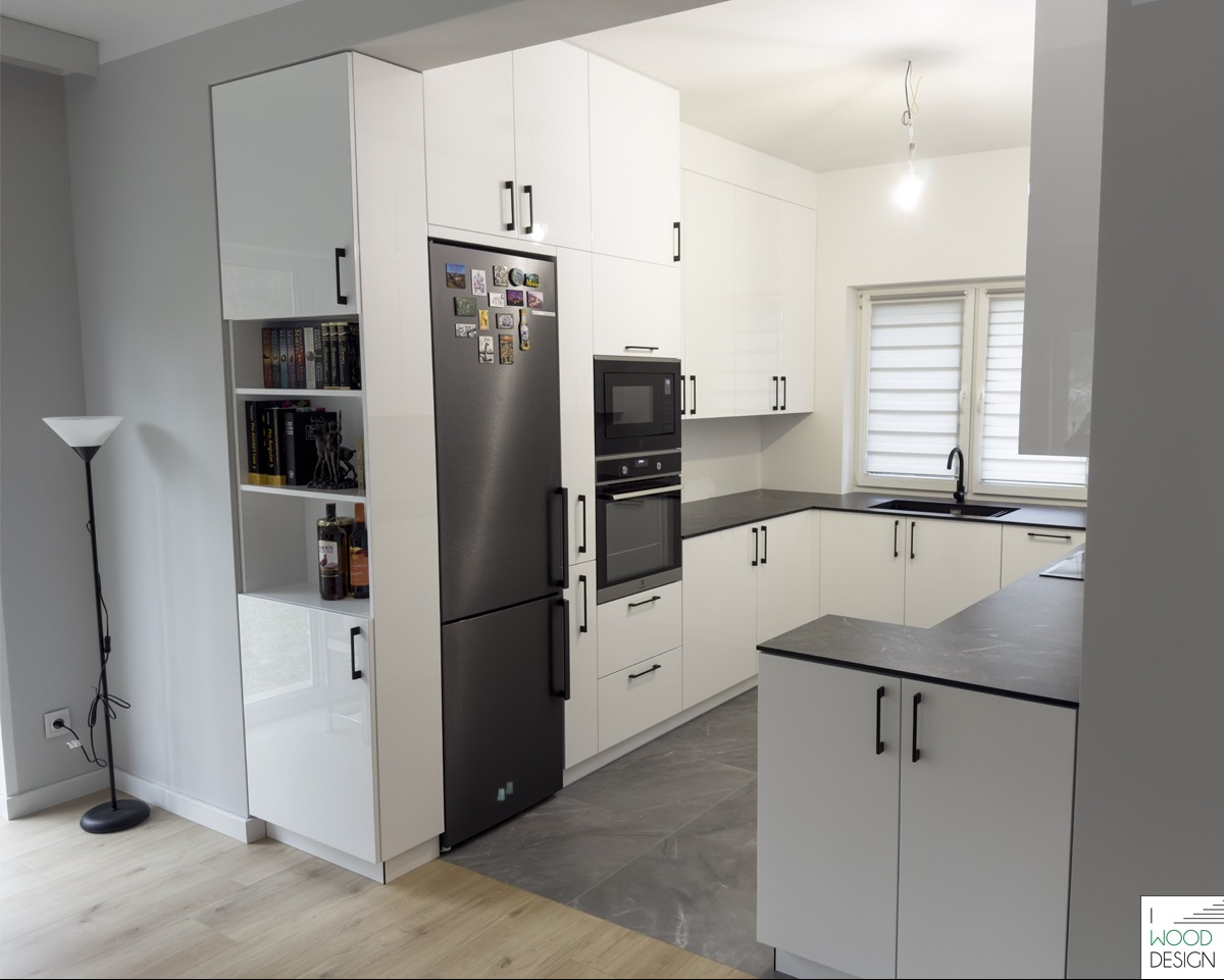 Nowoczesna kuchnia na wymiar z białymi, lakierowanymi frontami i czarnymi blatami. Widoczna zabudowa z lodówką i piekarnikiem oraz oświetlenie.