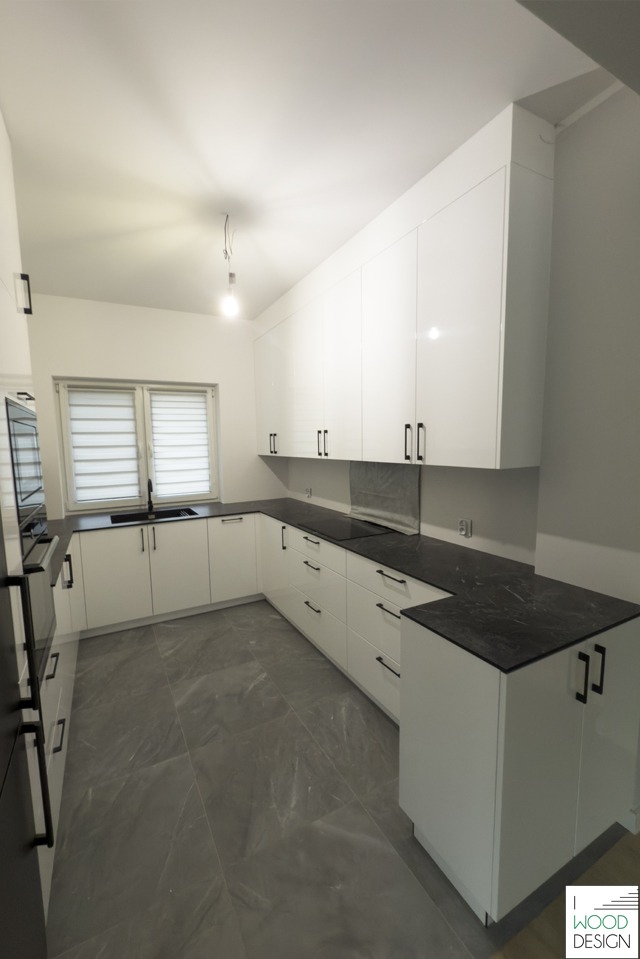 Biała, nowoczesna kuchnia z czarnym blatem i minimalistycznymi uchwytami. Widok na okno z żaluzjami i szarą podłogę. Oświetlenie sufitowe.