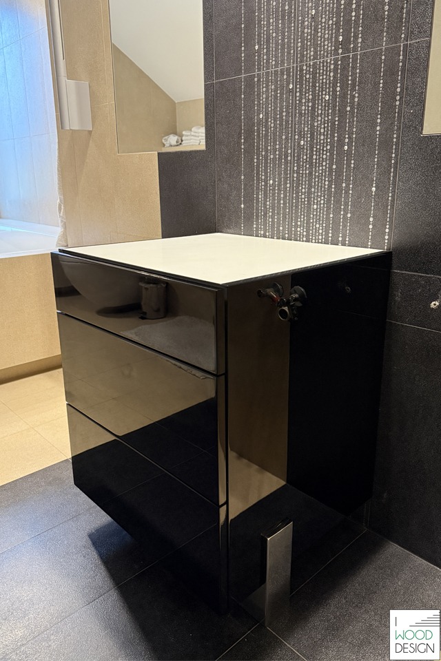 Czarna, lakierowana szafka łazienkowa z trzema szufladami i białym blatem, na tle ciemnych płytek z dekoracyjnym wzorem. Widoczne elementy instalacji wodnej.