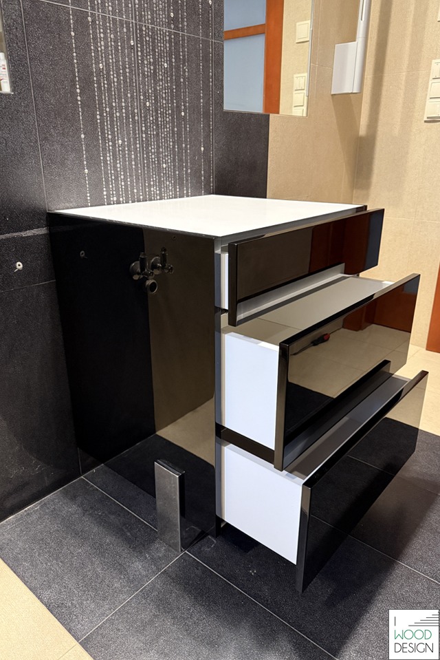 Nowoczesna szafka łazienkowa z trzema szufladami, czarno-biała, z połyskującymi frontami. Widoczne elementy instalacji wodnej i fragment ściany z dekoracyjnymi płytkami.