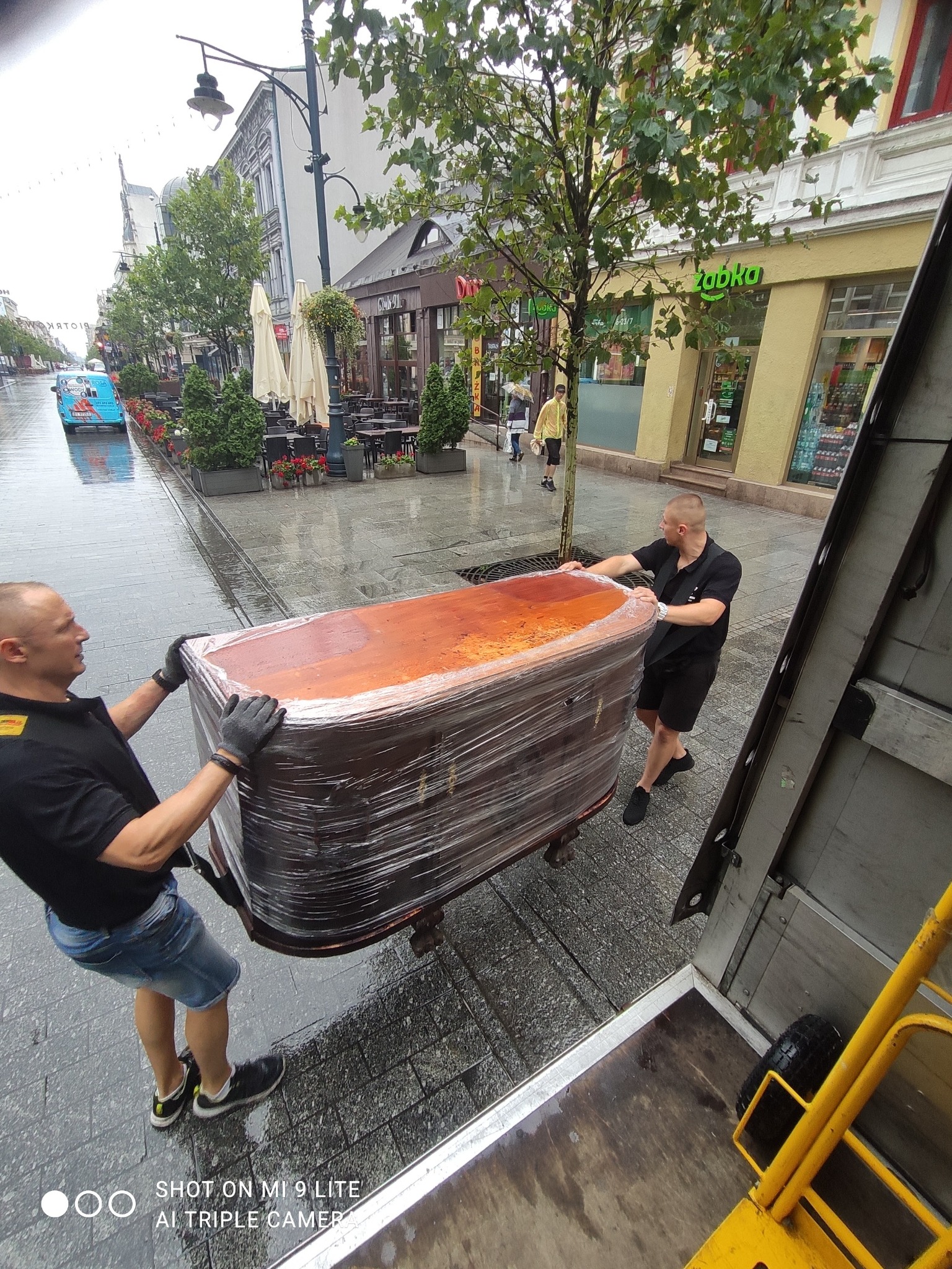 Dwóch mężczyzn w czarnych koszulkach i rękawicach transportuje owinięte folią biurko na chodniku w deszczowy dzień, widoczny fragment windy transportowej i miejski krajobraz z restauracjami...