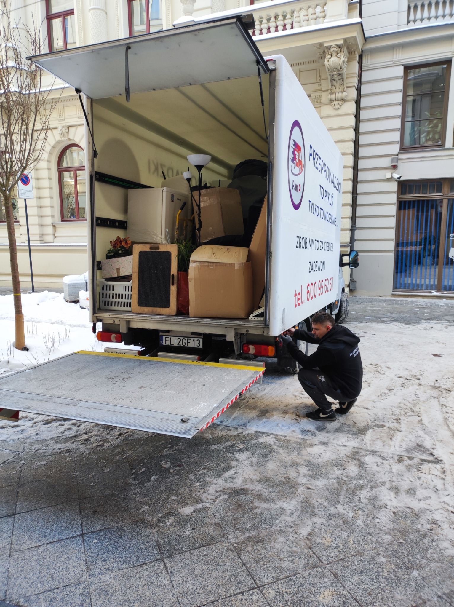 Otwarta paka busa wypełniona kartonami, meblami i lampą, pracownik w czarnej bluzie reguluje rampę załadunkową na tle miejskiej architektury i zimowego chodnika.
