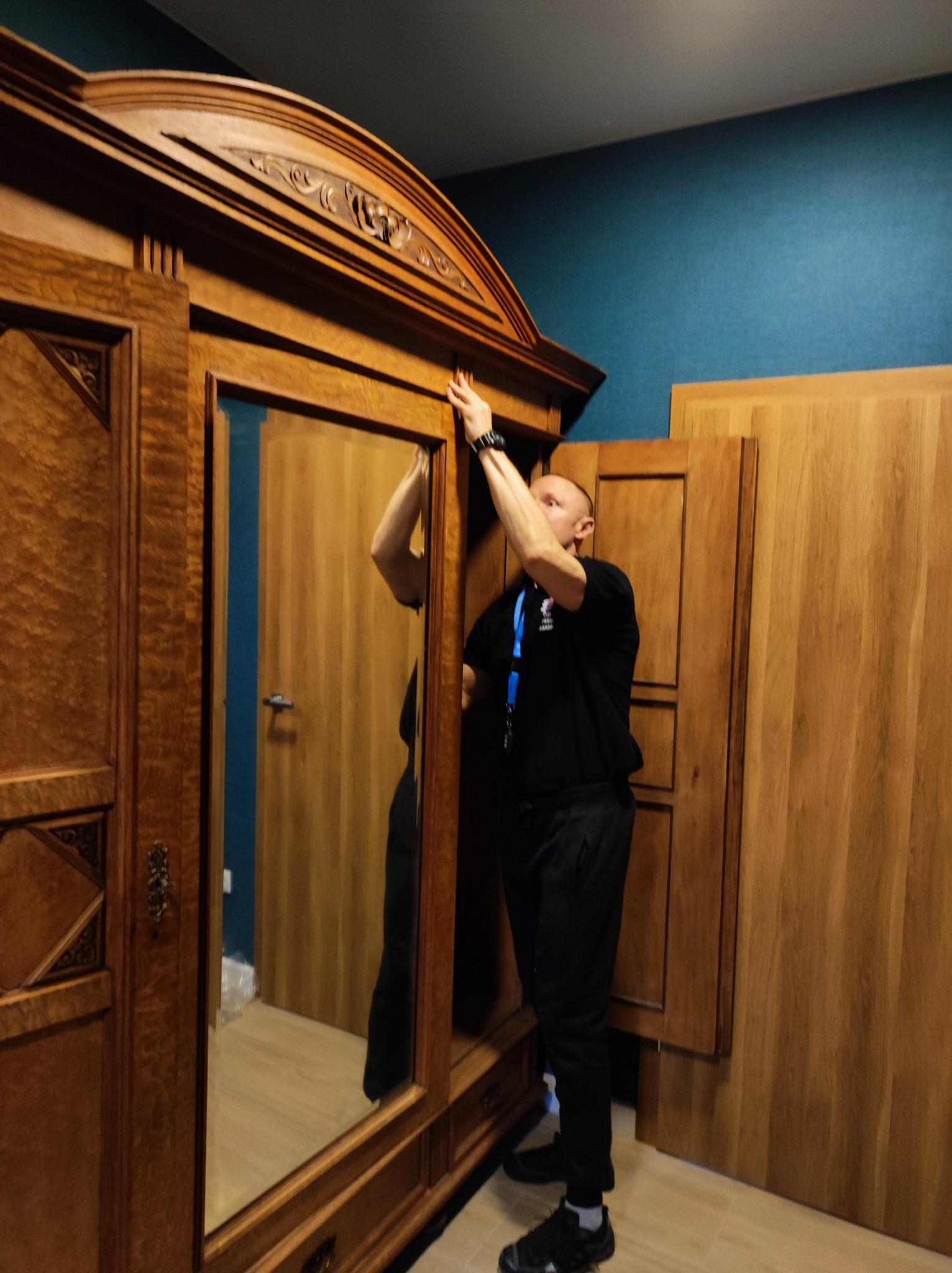 Mężczyzna w czarnym stroju, z identyfikatorem na szyi, podtrzymuje górną część dużej, ozdobnej, drewnianej szafy z lustrem podczas montażu lub przenoszenia.