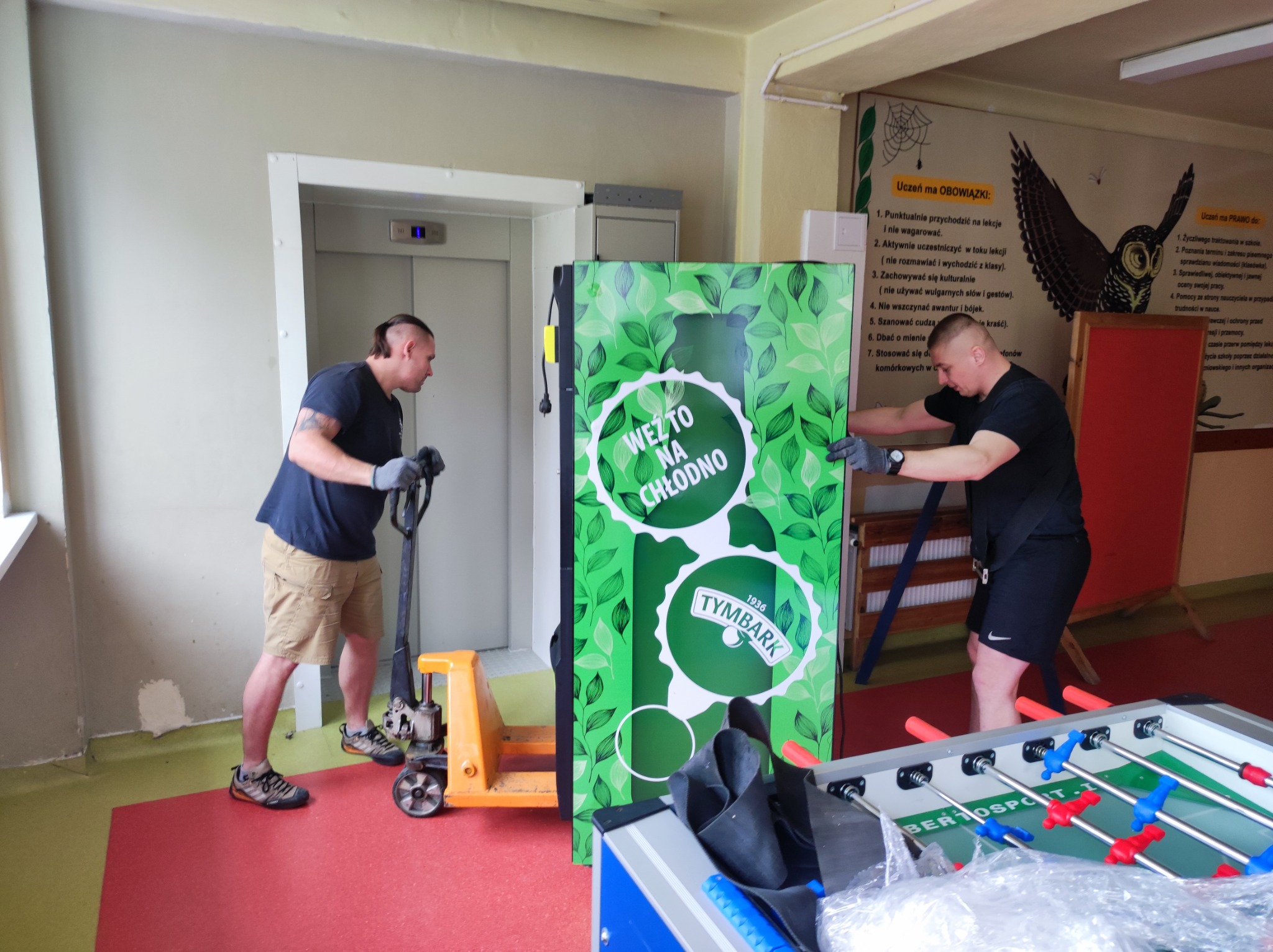 Dwóch mężczyzn w Łodzi transportuje na paleciaku automat z napojami Tymbark wewnątrz budynku, prawdopodobnie szkoły, obok windy i stołu do piłkarzyków.