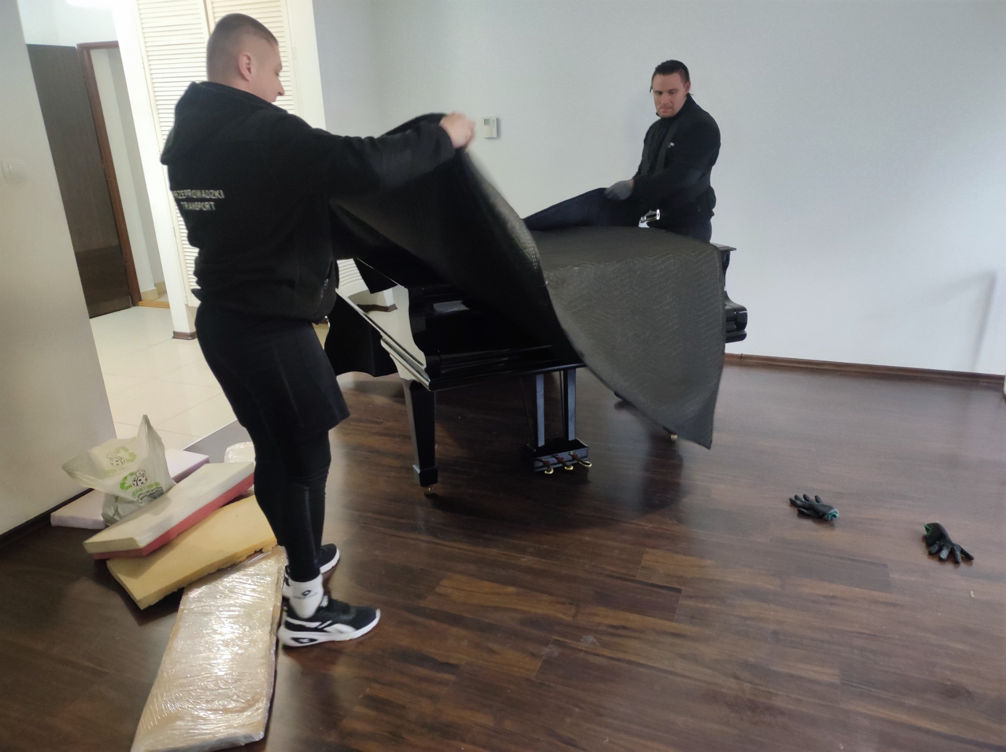 Dwóch mężczyzn w czarnych uniformach owija czarne pianino ochronnym kocem podczas przeprowadzki. Na podłodze leżą rękawice robocze i materiały zabezpieczające.