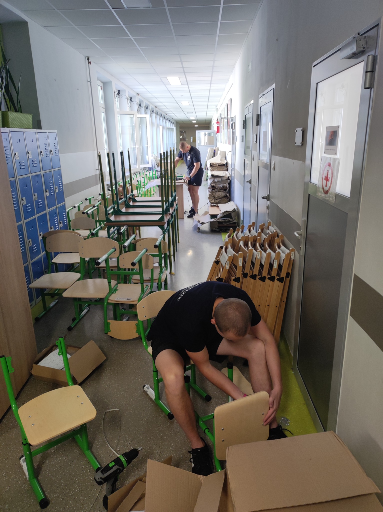 Dwaj mężczyźni składają nowe krzesła szkolne na korytarzu z niebieskimi szafkami i stosem złożonych krzeseł w tle, widoczne kartony i wkrętarka.