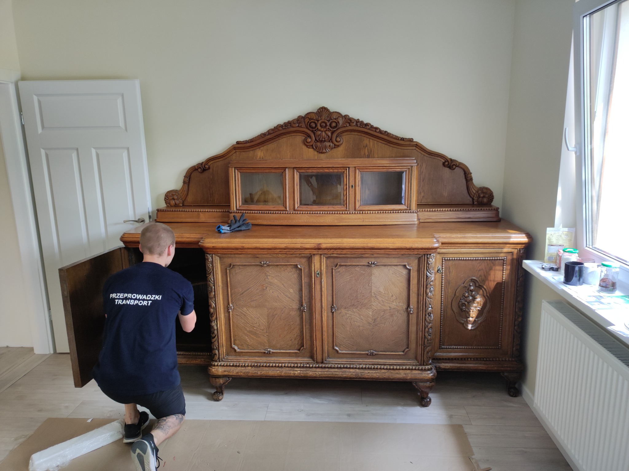 Pracownik firmy przeprowadzkowej w koszulce z napisem 'Przeprowadzki Transport' demontuje zabytkowy, zdobiony kredens z drewnianymi drzwiczkami i przeszkleniami.