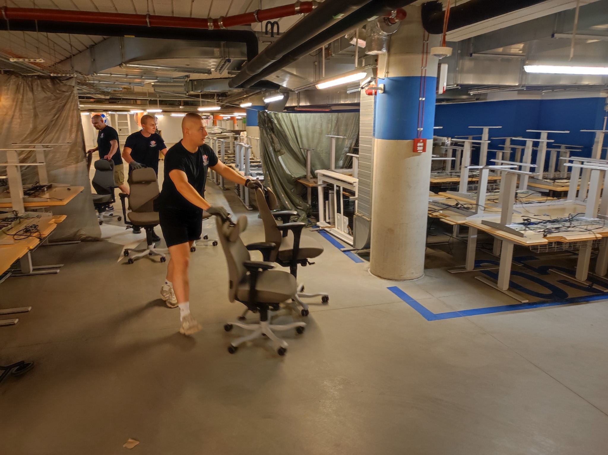 Mężczyźni w czarnych koszulkach i rękawicach transportują krzesła biurowe w przestrzeni biurowej z demontowanymi biurkami, widoczne instalacje pod sufitem.