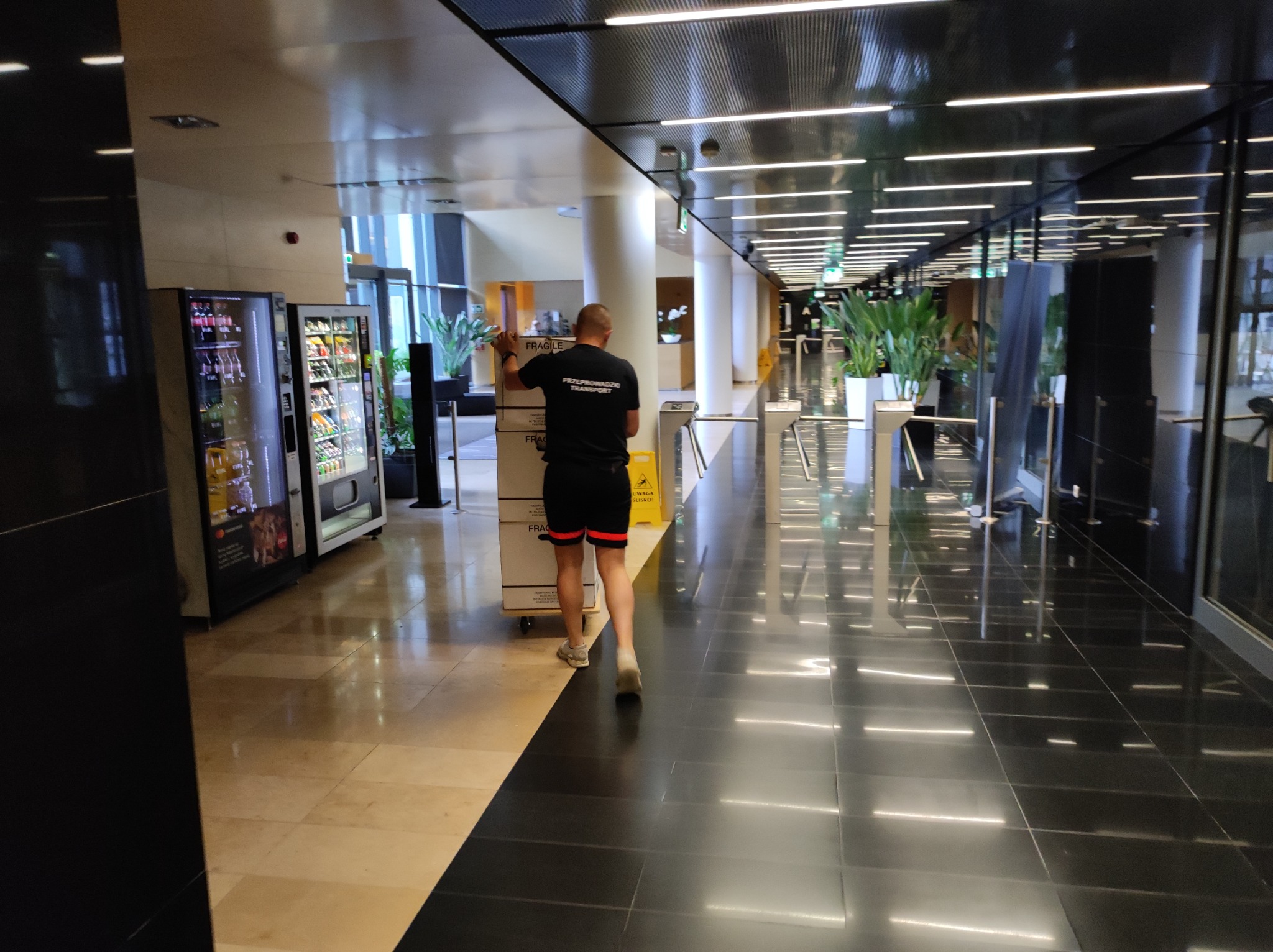 Pracownik w czarnej koszulce z napisem 'Przeprowadzki Transport' pcha wózek z kartonami oznaczonymi jako 'Fragile' w nowoczesnym biurowcu z automatami z napojami w tle.