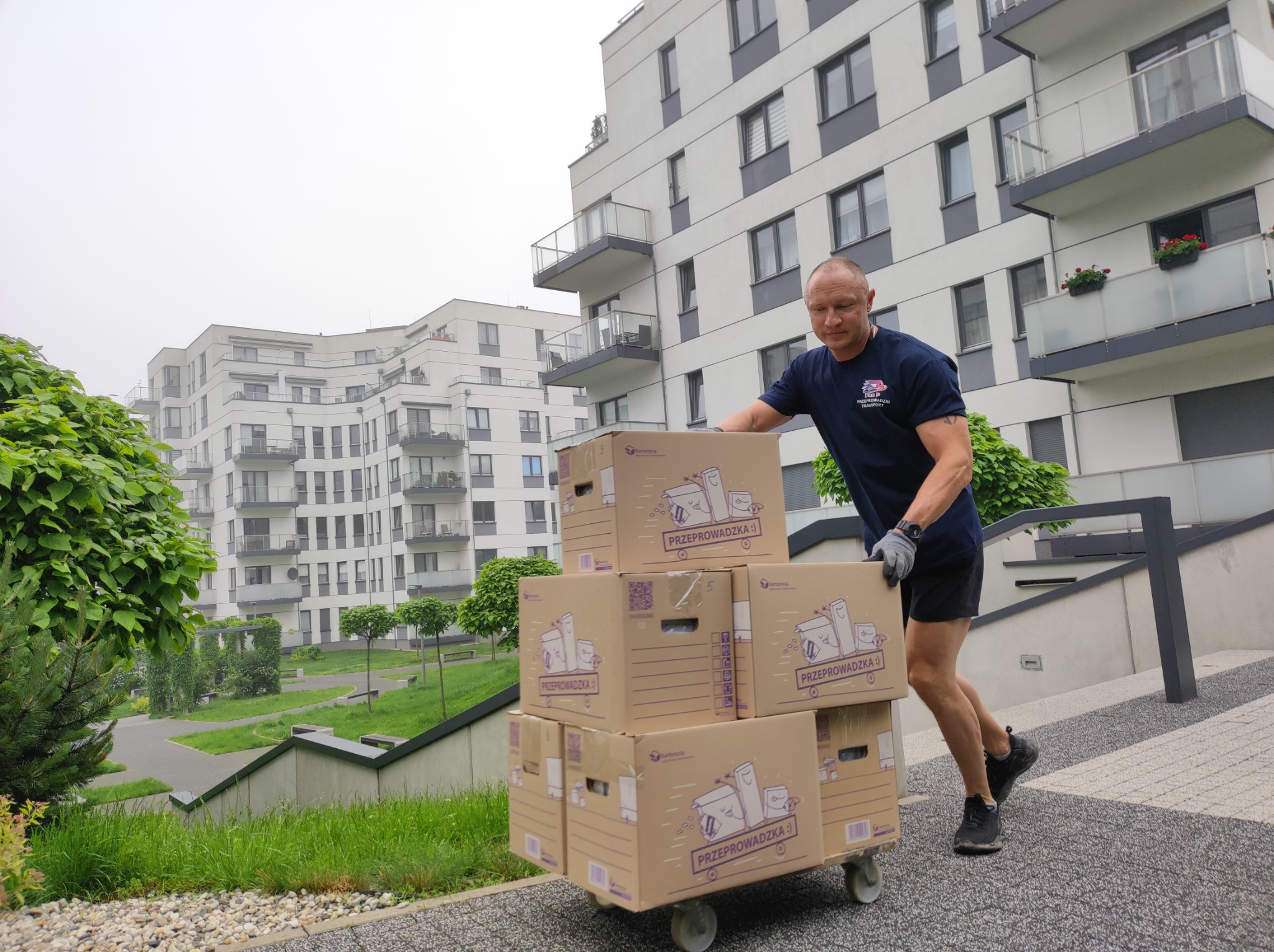 Mężczyzna w rękawicach pcha wózek z kartonami oznaczonymi napisem 'Przeprowadzka' na tle nowoczesnego osiedla.
