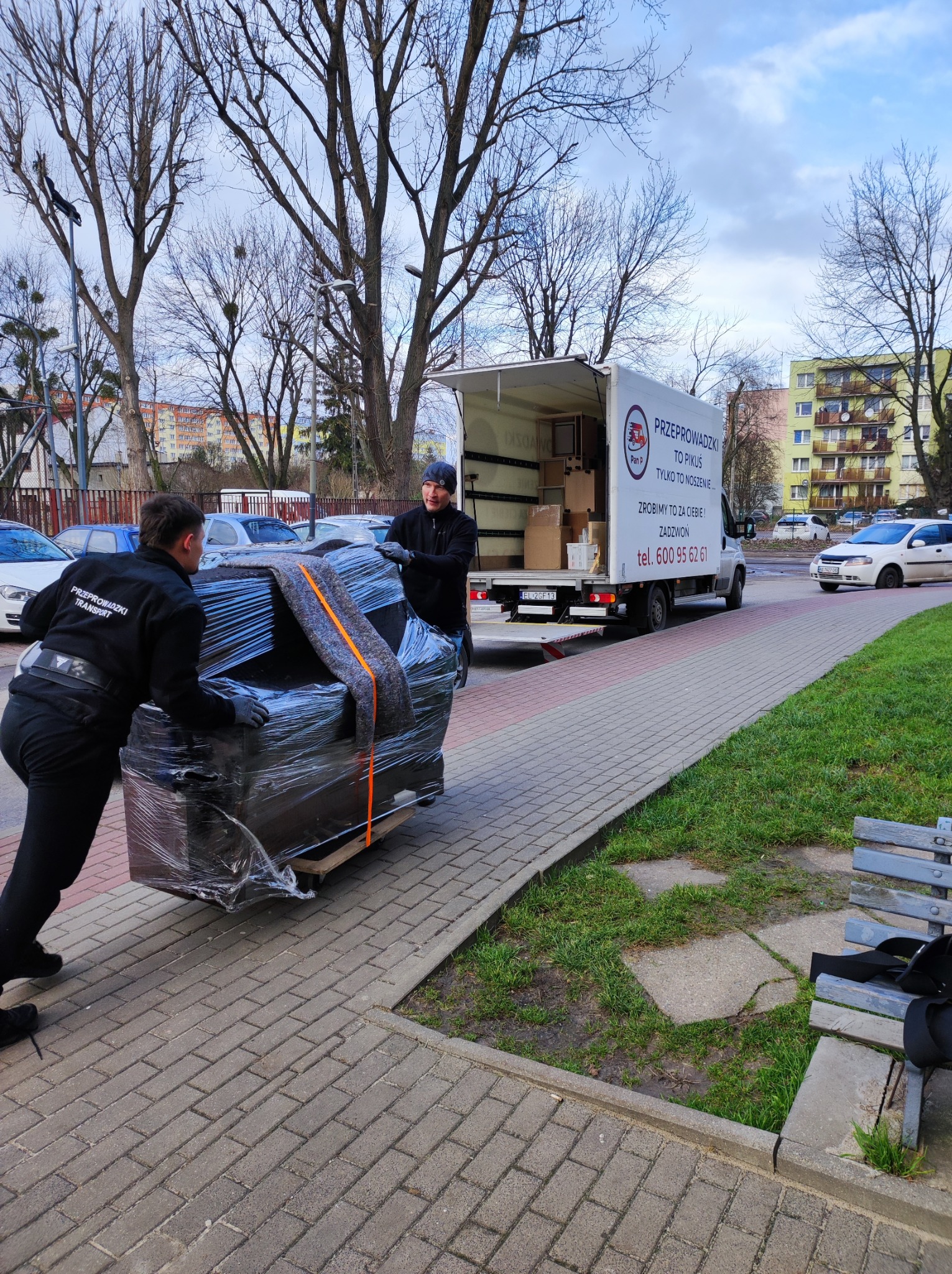 Dwóch mężczyzn w czarnych uniformach transportuje owinięte folią pianino na wózku po chodniku w kierunku białej furgonetki z napisem 'Przeprowadzki to Pikuś' na tle budynków mieszkalnych i drzew...