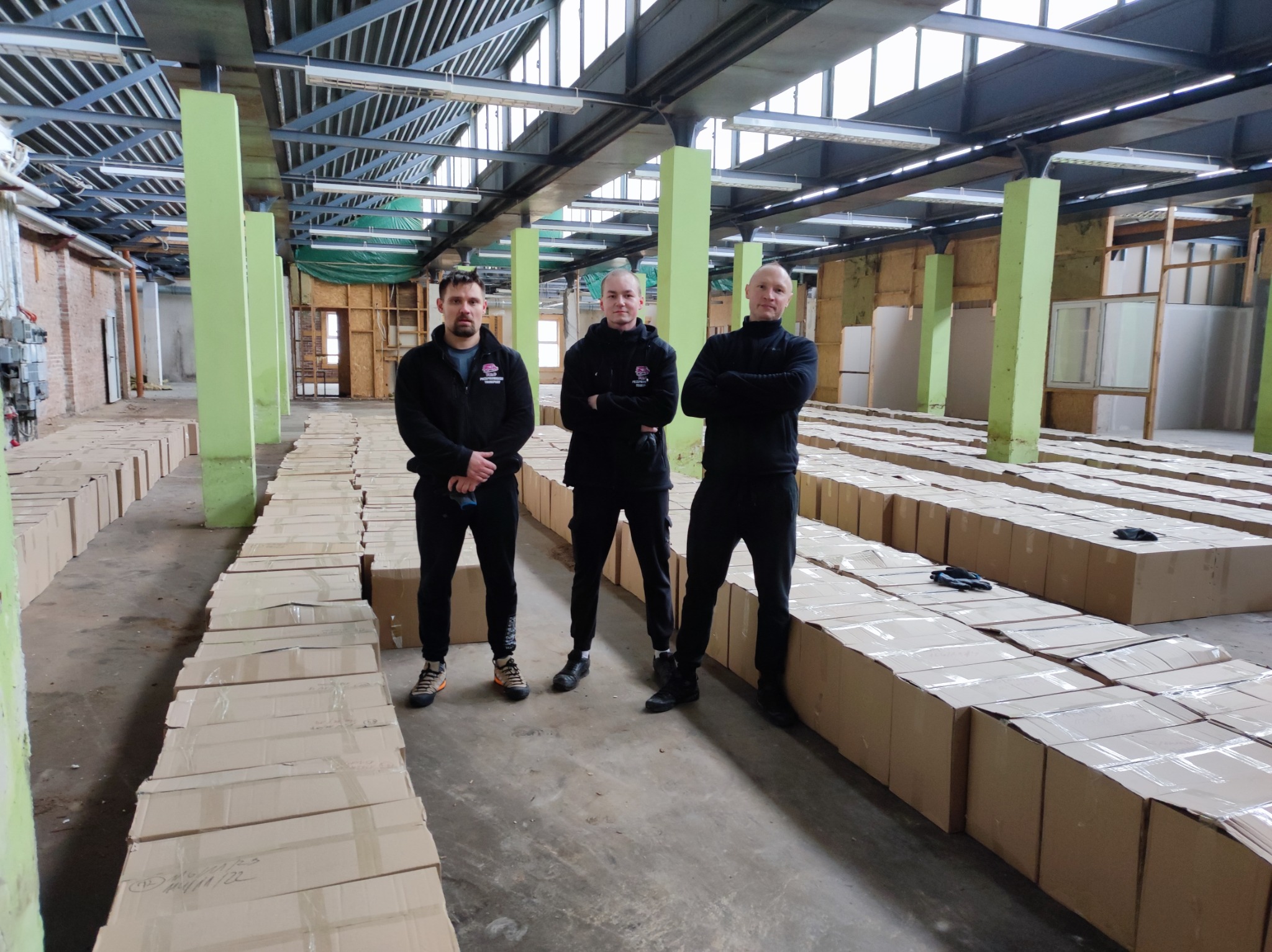 Trzech pracowników firmy transportowej stoi na hali magazynowej pomiędzy rzędami kartonowych pudeł zabezpieczonych folią, gotowych do przewozu. Pudełka ułożone w długie rzędy, widoczne zielone...