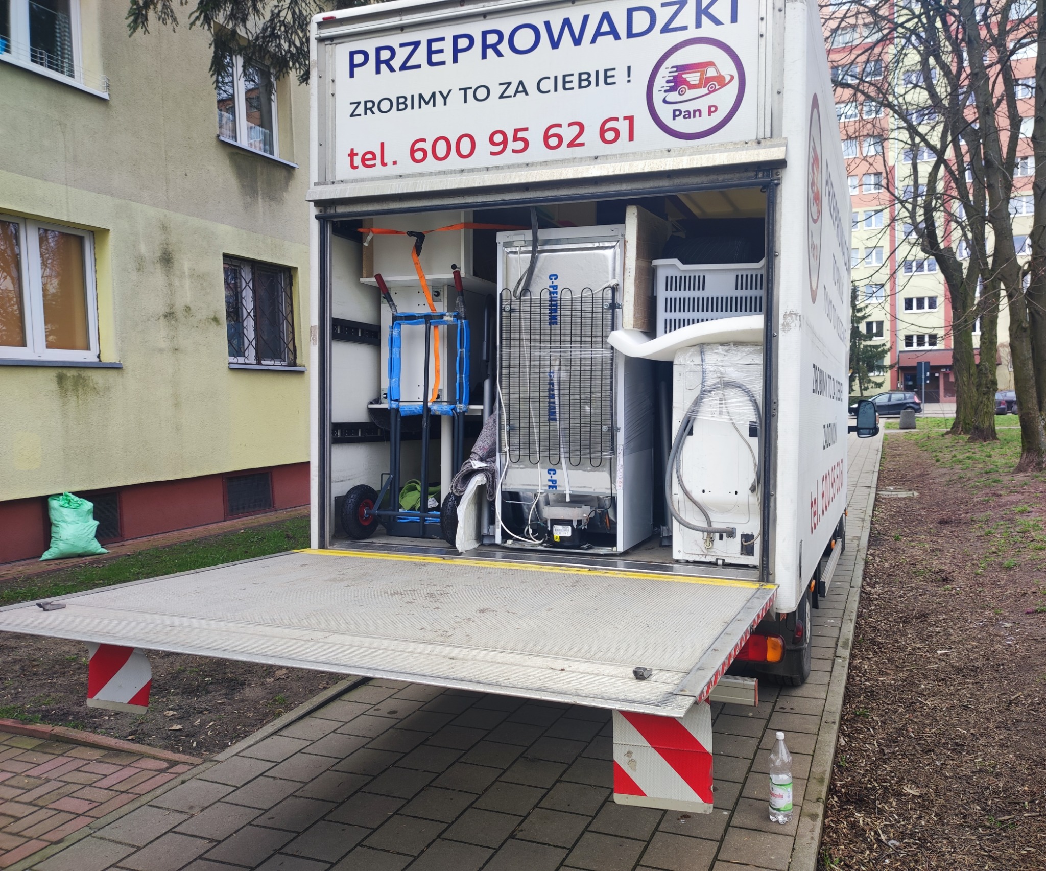 Otwarta paka samochodu dostawczego firmy przeprowadzkowej z widocznym sprzętem AGD (lodówka i pralka) zabezpieczonym do transportu, w tle budynek mieszkalny.