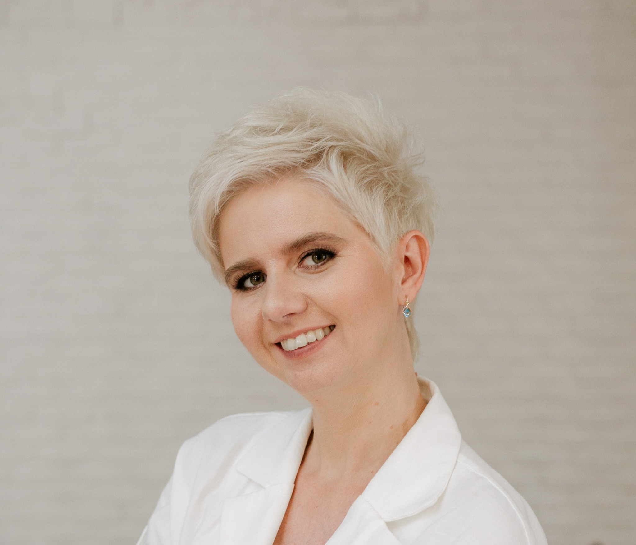 Portret uśmiechniętej kobiety w białym żakiecie, z krótkimi blond włosami i subtelnym makijażem, pozującej na jasnym tle.