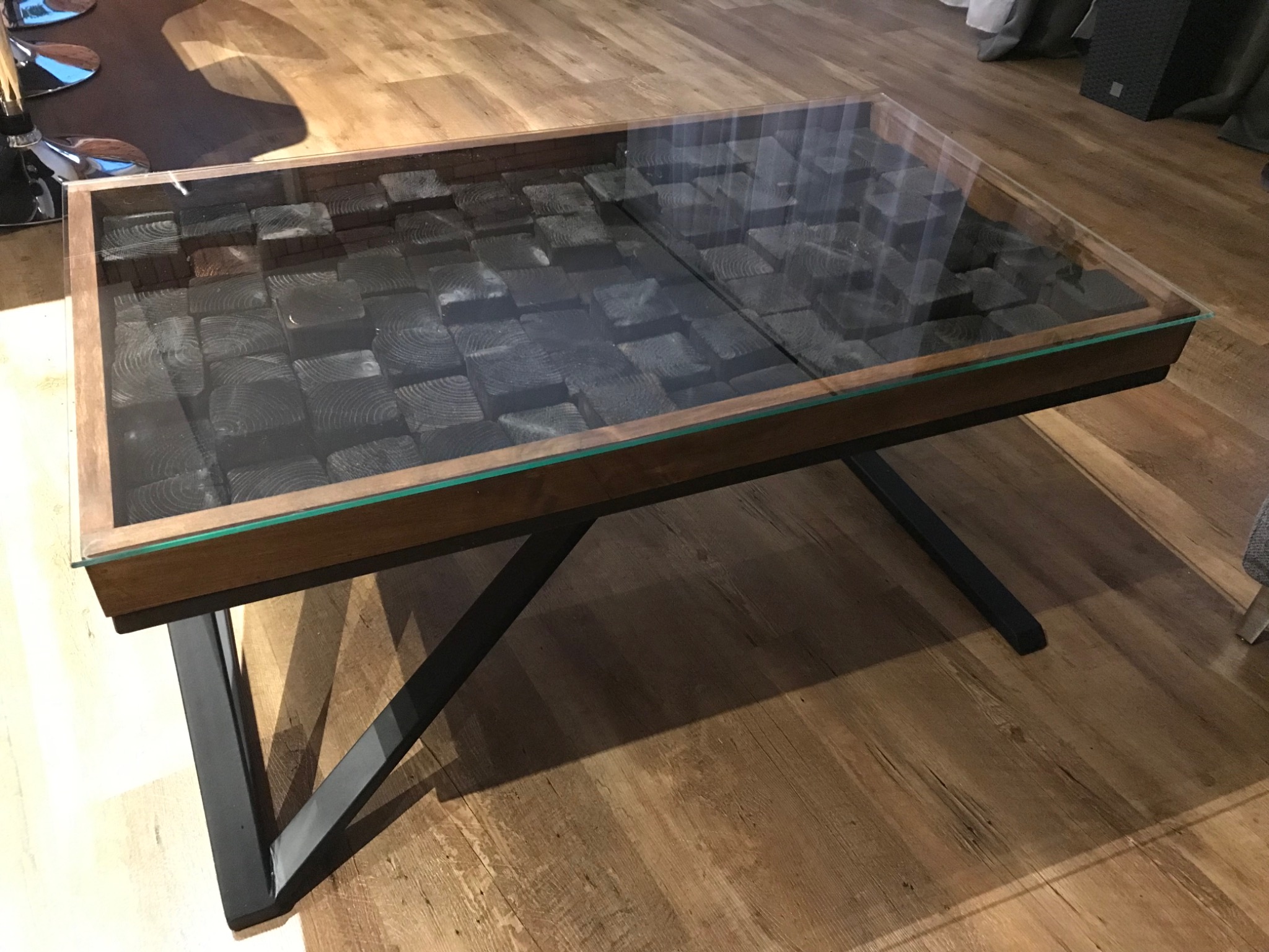 Stolik kawowy z drewnianym blatem wypełnionym czarnymi kostkami drewna pod szklaną powierzchnią, wsparty na metalowych nogach w kształcie litery V, na tle drewnianej podłogi.