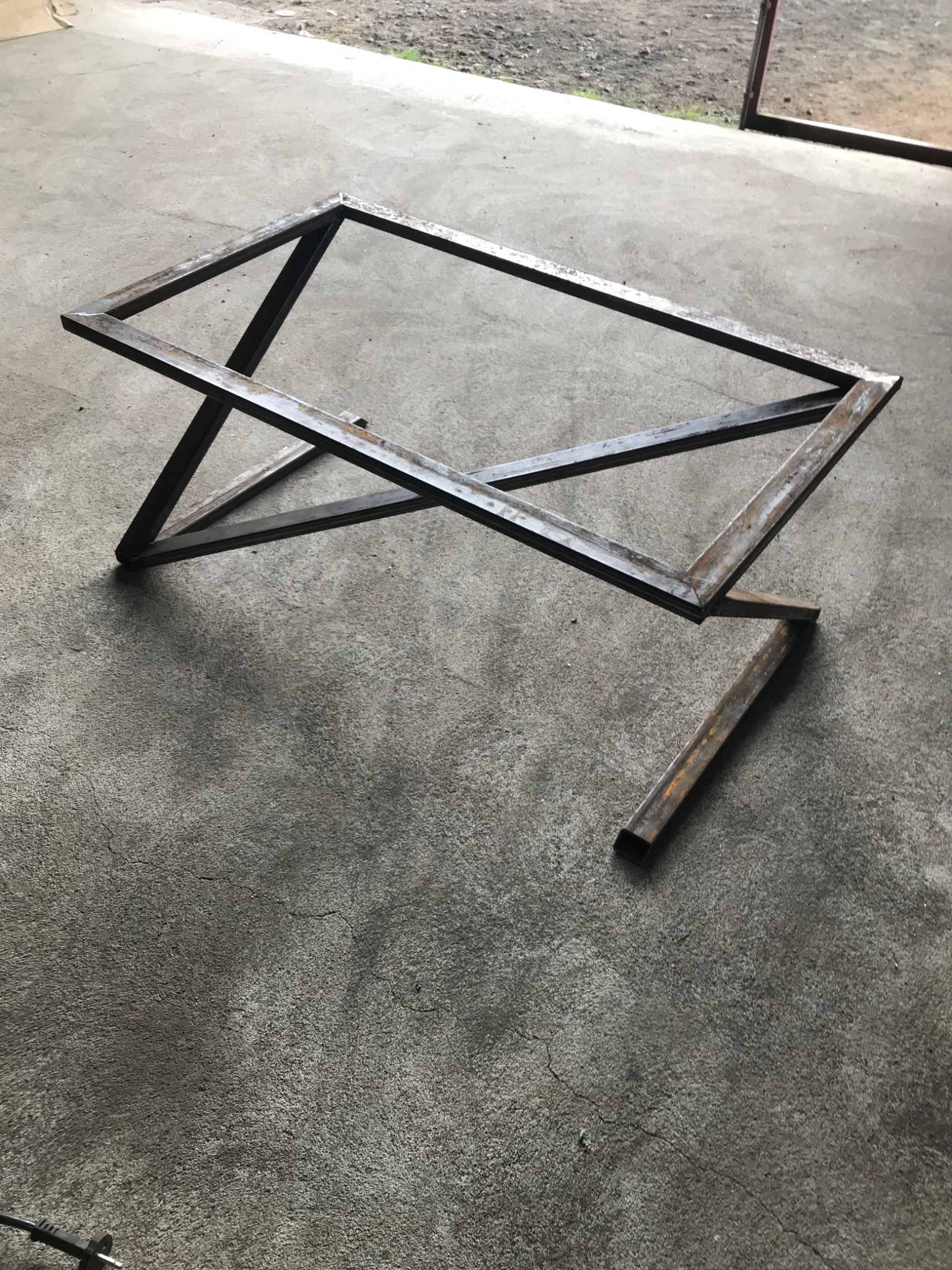 Metalowa konstrukcja stolika kawowego z profili kwadratowych, surowe spawy, widoczne ślady korozji, fragment niezamocowanego elementu na betonowej posadzce.