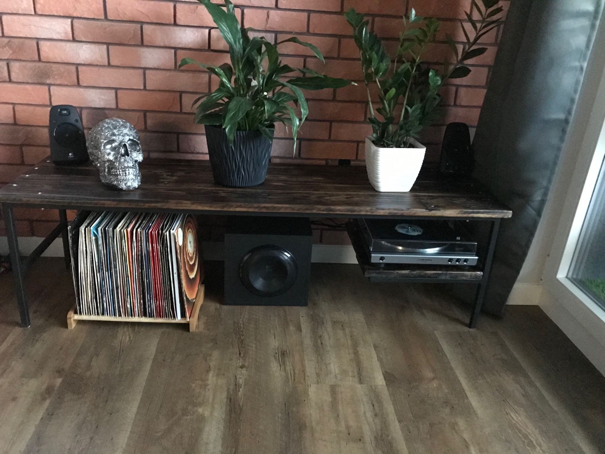 Ręcznie robiony stolik z drewna i metalu na którym stoją głośniki, dekoracyjna czaszka, dwie rośliny w doniczkach, pod spodem gramofon, subwoofer i kolekcja płyt winylowych w drewnianym stojaku...