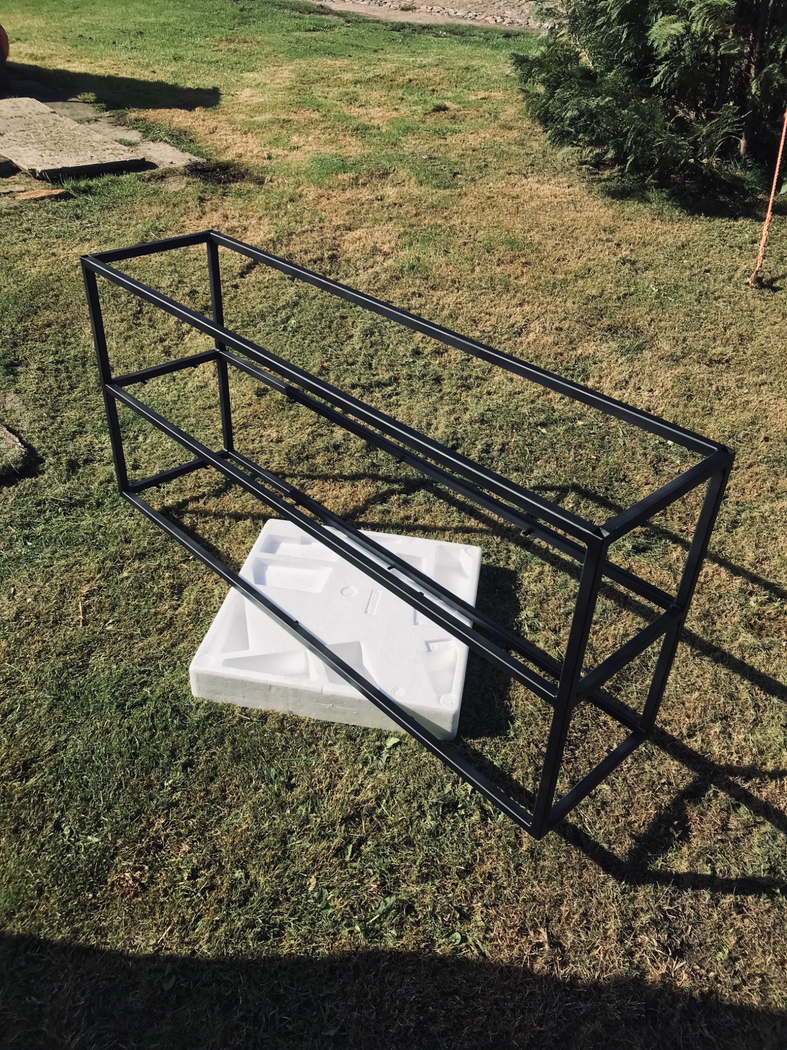 Czarna, metalowa rama regału spawana w Szczecinie, ustawiona na białym styropianie na trawie w ogrodzie.