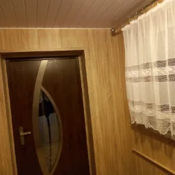 Wnętrze przedpokoju wykończone panelami imitującymi drewno, z ciemnobrązowymi drzwiami z szybą o owalnym kształcie, zasłonięte okno z białą, koronkową firanką i widoczne pantofle pod ścianą.