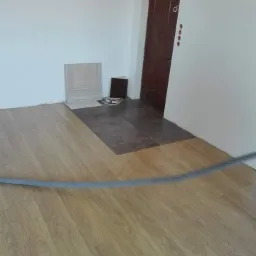 Fragment pokoju z ułożoną podłogą z paneli imitujących drewno, z widocznym progiem z płytek w odcieniach brązu, przy ścianie leżą próbki paneli i płytek, widoczna brązowa drewniana ościeżnica drzwi