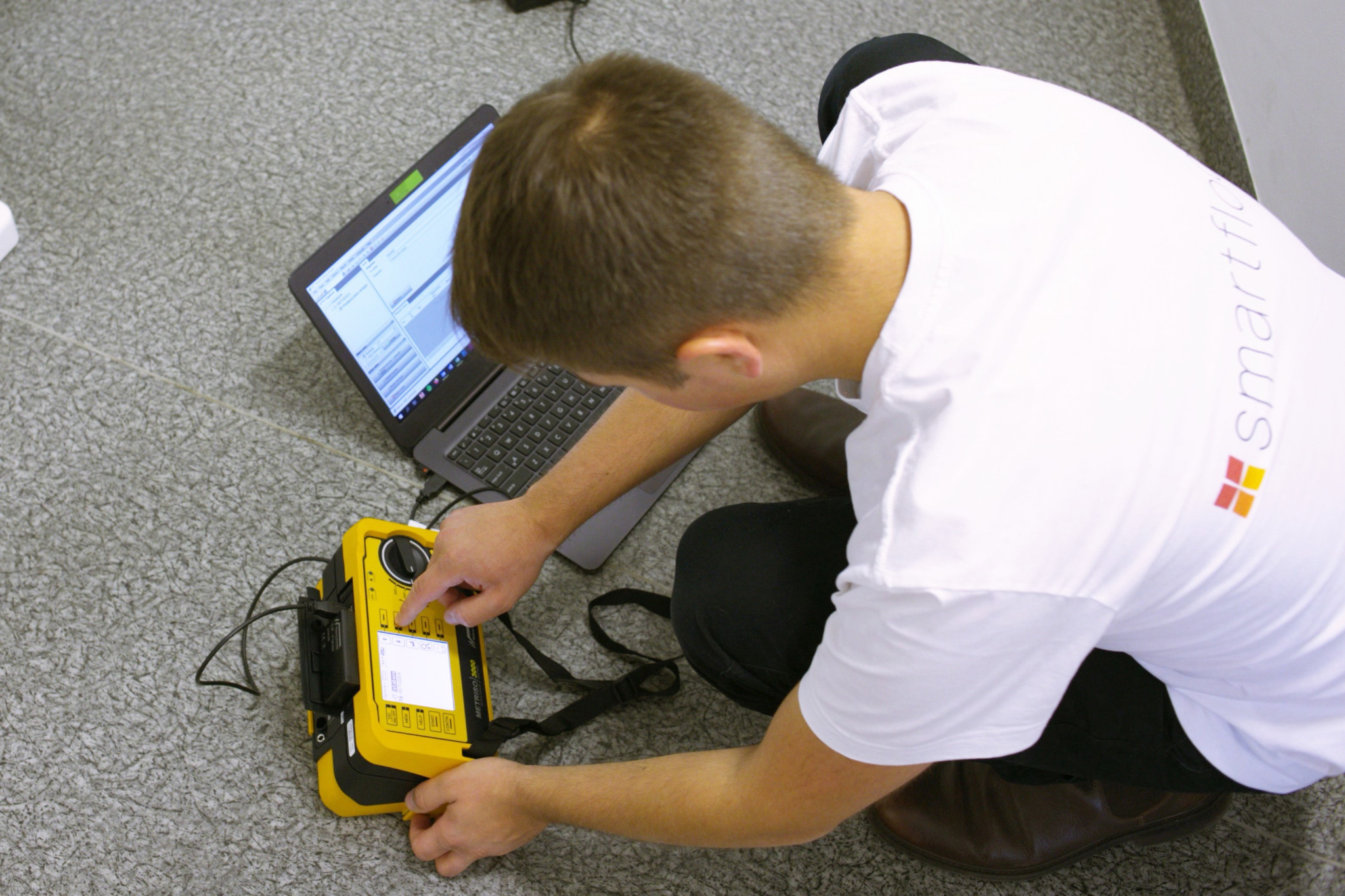 Osoba w białej koszulce z logo firmy obsługuje żółty miernik elektryczny Metrel Eurotest 3000, podłączony do laptopa, na tle szarej wykładziny podłogowej.