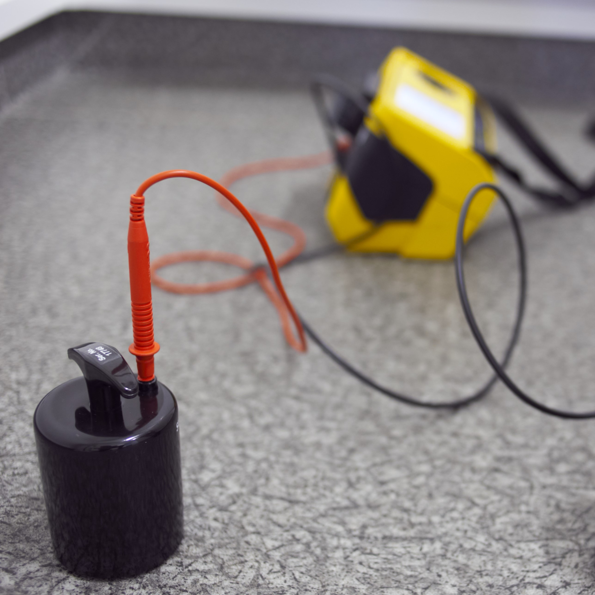 Czarny cylinder z podłączonym czerwonym przewodem pomiarowym, w tle żółty miernik na szarej powierzchni.