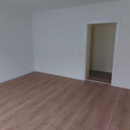 AIG-Company - Puste, pomalowane na biało pomieszczenie z panelami podłogowymi w odcieniach brązu, widoczne gniazdka elektryczne i listwy przypodłogowe, z otwartym przejściem do kolejnego pomieszczenia.