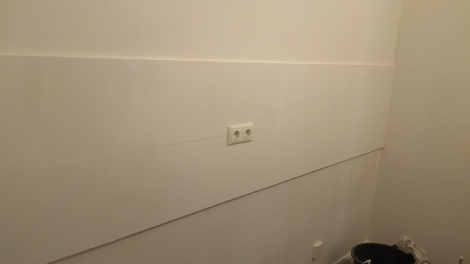 Biała ściana z zamontowanym gniazdkiem elektrycznym, widoczne łączenie paneli ściennych, w rogu fragment wiadra.