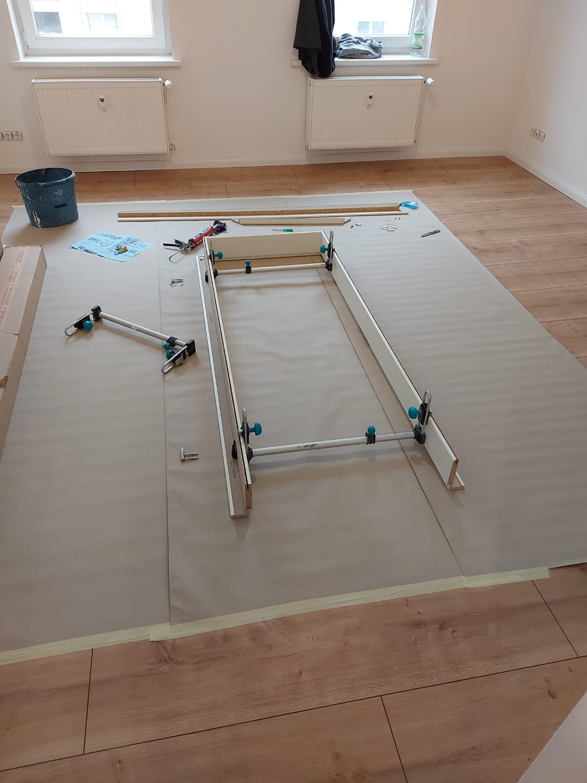 Przygotowanie ramy drzwiowej na podłodze zabezpieczonej folią malarską, widoczne narzędzia montażowe, ściski stolarskie, listwy przypodłogowe.