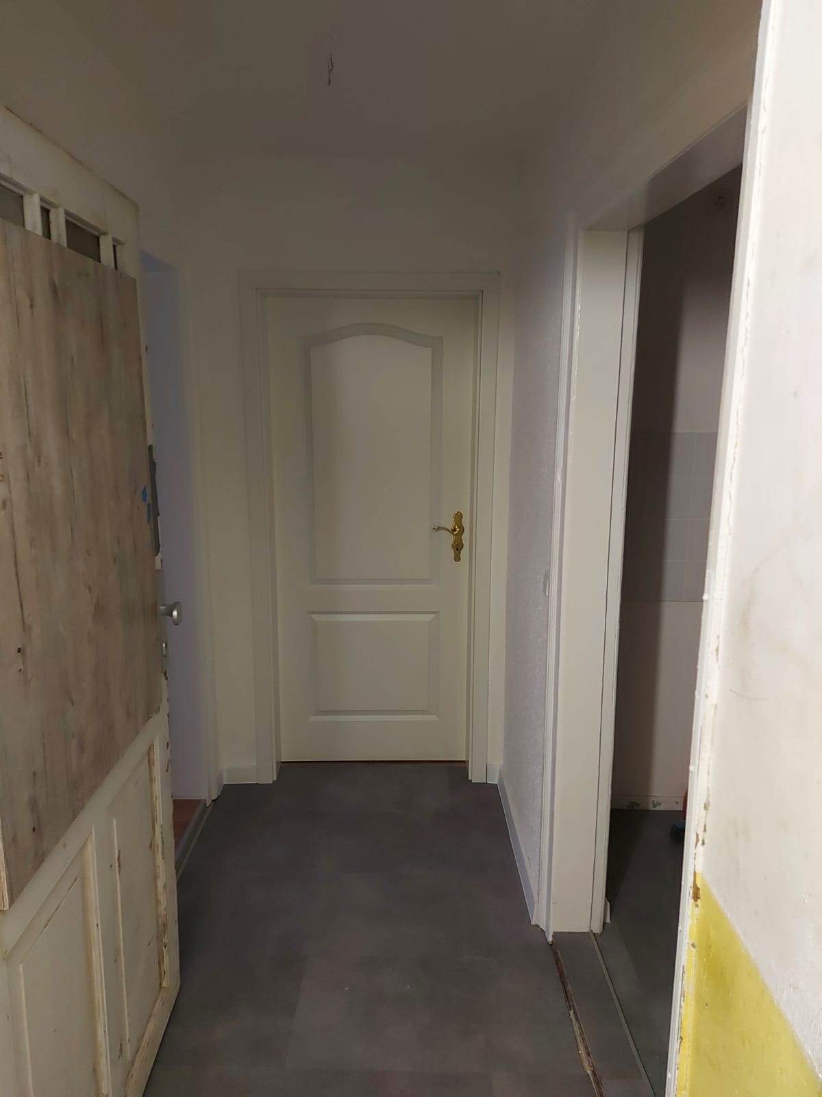Wnętrze korytarza z dwiema białymi drzwiami, szarą podłogą i widoczną ścianą z żółtymi plamami, sugerującymi prace wykończeniowe.