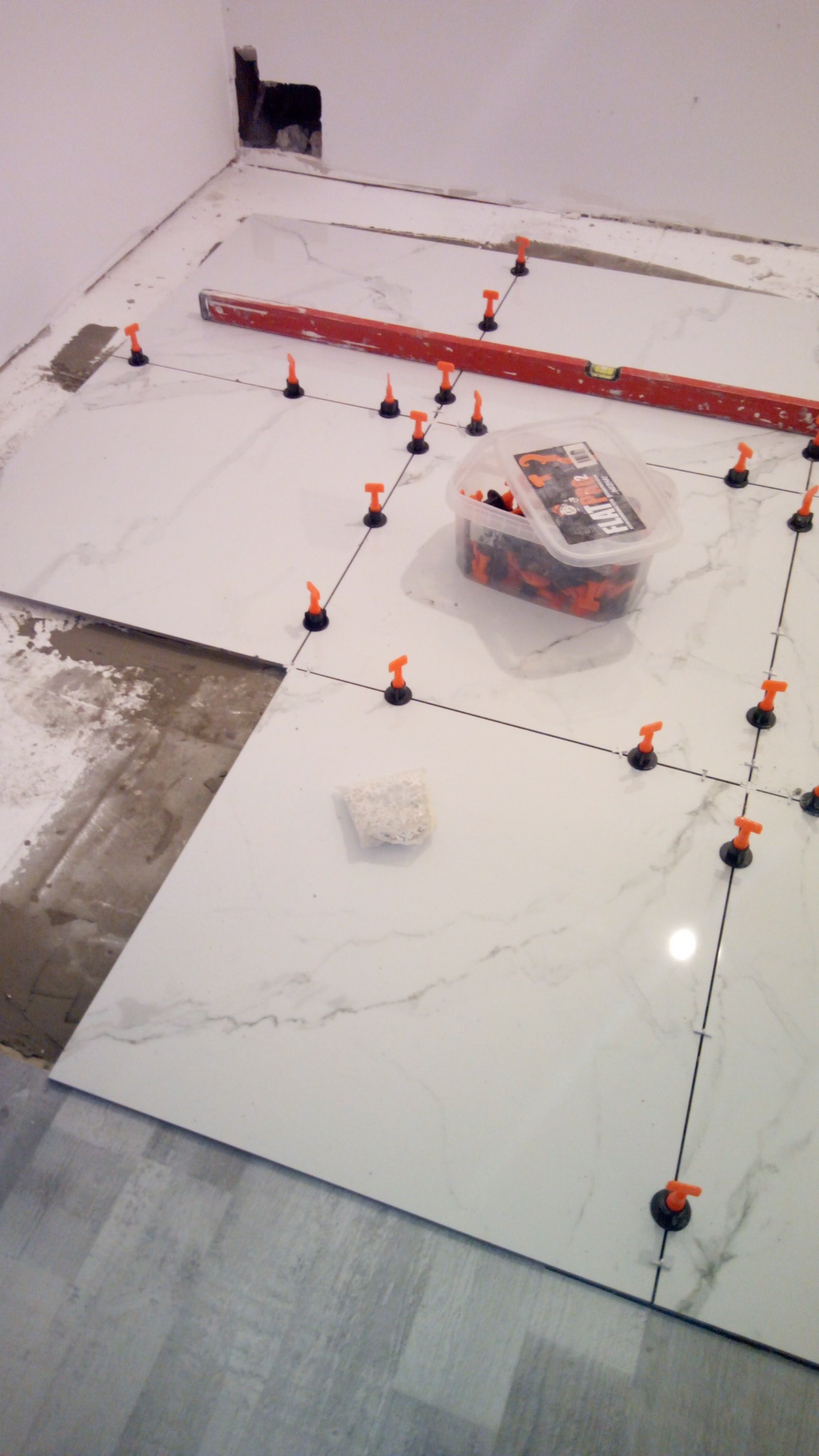 Układanie białych płytek podłogowych z imitacją marmuru, z zastosowaniem systemu poziomowania płytek (klipsy i kliny) oraz poziomicy, widoczne resztki kleju i fragment starej podłogi.