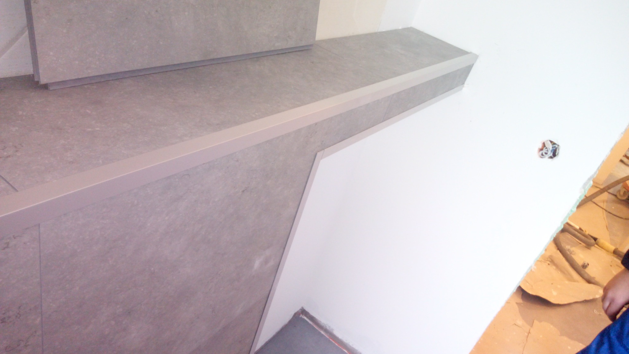 Wykończenie schodów płytkami ceramicznymi w odcieniach szarości z aluminiowymi listwami krawędziowymi, widoczny fragment ściany z przygotowanym otworem na gniazdko elektryczne, podłoga...